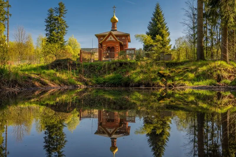 Na zdjęciu widać drewnianą cerkiew z kopulastymi dachami, otoczoną przez drzewa, z elegancką fasadą i prostą, ale dostojną architekturą, która emanuje spokojem i tradycją.