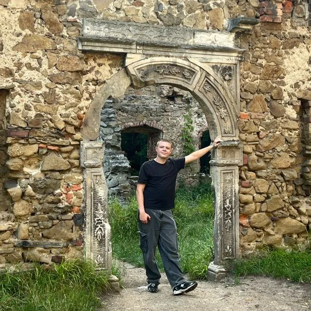 Oliwier Trager stojí ve vchodu obklopeném ozdobným kamenným portálem a rukou ukazuje na detail zničené zdi bývalé budovy.