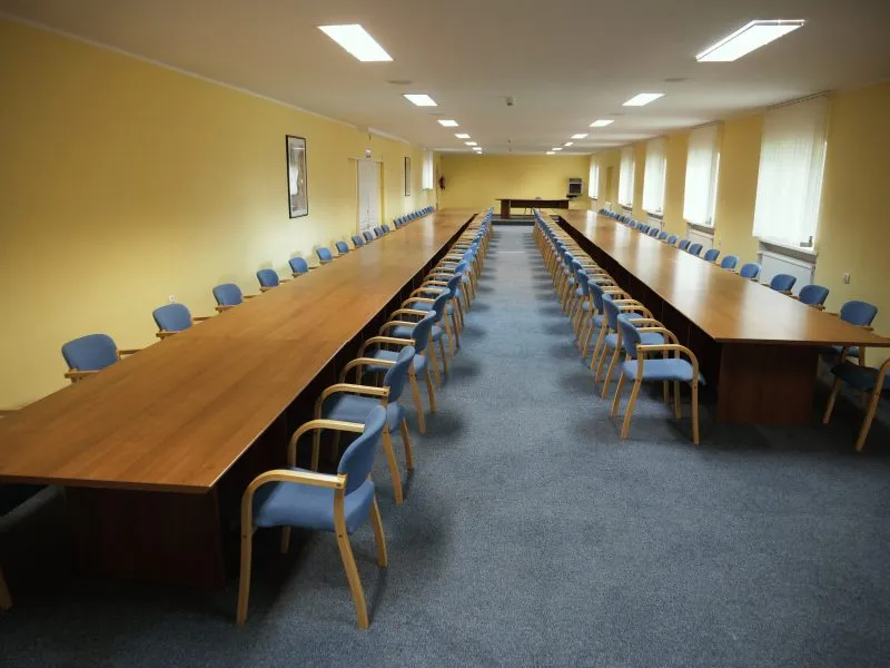 Duża, jasna salę konferencyjną z długimi stołami ustawionymi w dwóch rzędach i wieloma niebieskimi krzesłami. Pomieszczenie jest przestronne, równomiernie oświetlone sufitowymi lampami i przygotowane do spotkań lub szkoleń.