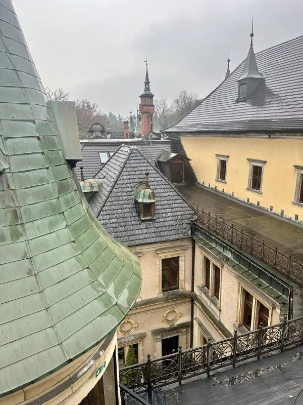 Výhled z Jennyiny věže na zámku Kliczków zahrnuje zelené střechy, kamennou terasu a věže rezidence, v dálce se tyčí věž kostela.