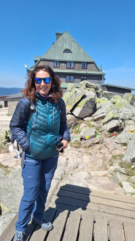 Agata Modzelewska pózuje v slunečních brýlích a turistickém oblečení na skalnatém vrcholu s horskou chatou v pozadí.