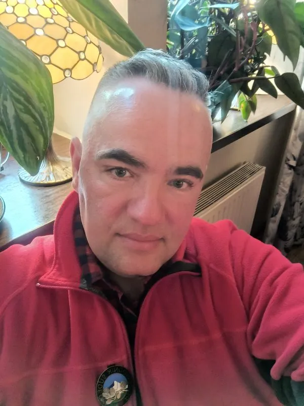 Andrzej Talarek si pořizuje selfie v červené fleecové bundě v interiéru s rostlinami, obrazy a dekorativní lampou v pozadí.