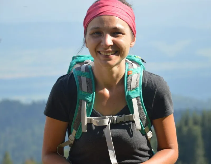 Marta Hamerla-Pater se usmívá v turistickém oblečení s růžovým šátkem na hlavě a batohem, stojící na pozadí rozlehlé horské krajiny.