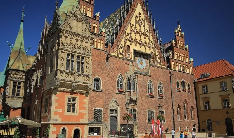 Bogato zdobiona gotycka fasada Starego Ratusza we Wrocławiu z zegarem astronomicznym, stromymi dachami i wieżyczkami lśni w ostrym słońcu.