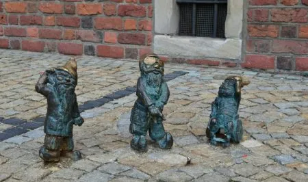 Trzy figurki wrocławskich krasnali stoją na brukowanym chodniku przy ceglanej ścianie, przedstawiając postacie z beczułką, długą brodą i małym zwierzęciem.