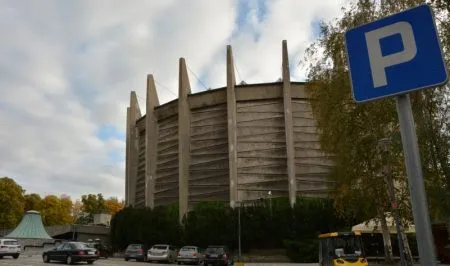 Okrągły żelbetowy gmach Panoramy Racławickiej we Wrocławiu wznosi się za parkingiem i drzewami, a na pierwszym planie stoi niebieski znak parkingowy.