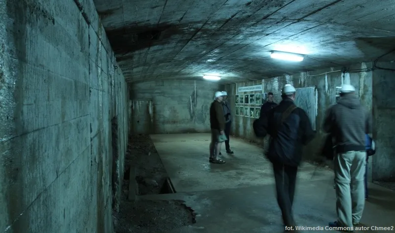 Turyści zwiedzają surowy betonowy korytarz kompleksu Osówka w Górach Sowich, idąc pod chłodnym światłem lamp wzdłuż masywnej ściany.
