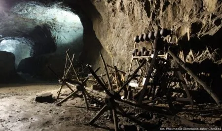 Mroczny, wykuty w skale tunel w Górach Sowich z dawnymi drewnianymi podporami i narzędziami, oświetlony chłodnym światłem wpadającym z bocznego korytarza.