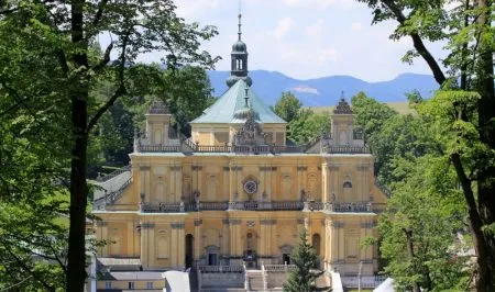 Monumentální barokní bazilika ve Vambeřicích se žlutou, bohatě zdobenou fasádou se tyčí nad korunami stromů na pozadí hor a zamračené oblohy.
