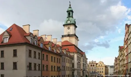 Řada historických měšťanských domů na náměstí v Jelení Hoře vede zrak k vysoké radniční věži se zelenou helmicí pod lehce zamračenou oblohou.