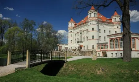 Biały Pałac Wojanów z czerwonymi dachami, narożnymi wieżami i ozdobną fasadą stoi w zadbanym parku za niewielkim mostkiem nad fosą.