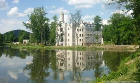 Jasna bryła pałacu w Karpnikach, z wieżami i odrestaurowanymi elewacjami, odbija się w spokojnej tafli stawu pośród drzew Doliny Pałaców i Ogrodów.