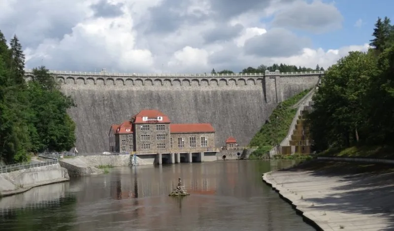 Zdjęcie przedstawiające kamienną tamę z niewielką elektrownią wodną u stóp