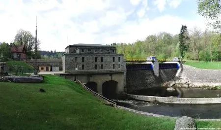 Elektrownia wodna we Wrzeszczynie stoi przy kamiennej zaporze i kaskadzie na Bobrze, otoczona trawiastymi skarpami, drzewami oraz dawną zabudową techniczną.