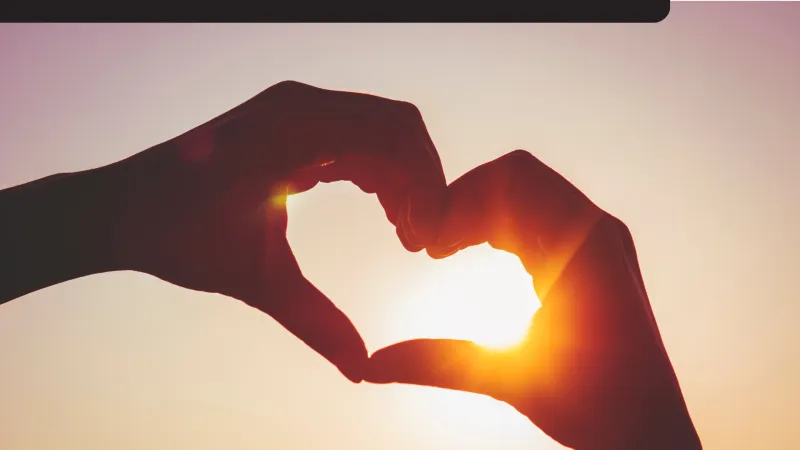 Deux mains forment un cœur autour du soleil couchant, créant un cadre chaleureux et romantique sur fond de ciel vespéral aux teintes douces.