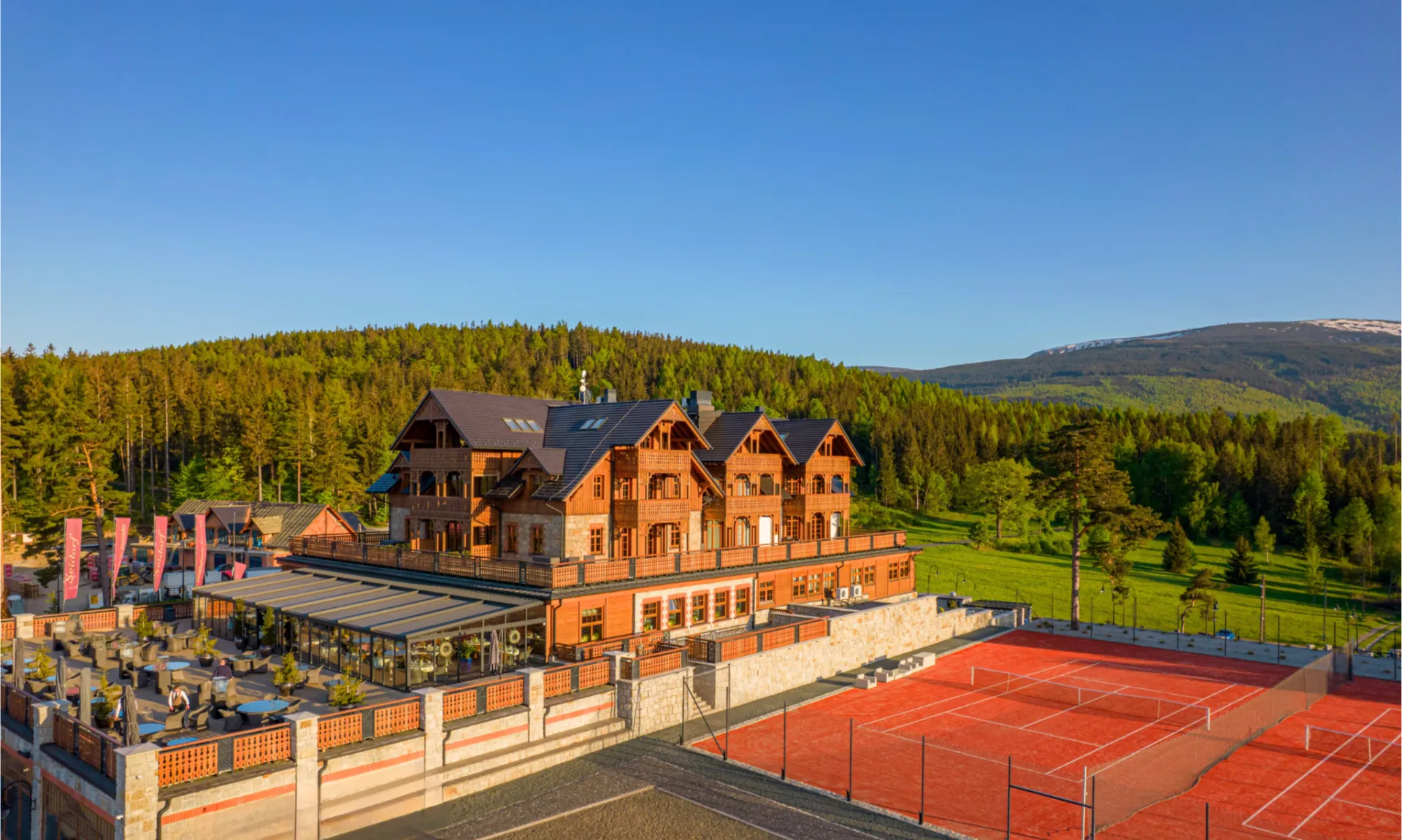 Elegancki hotel w stylu górskiego kurortu, położony wśród zielonych lasów u podnóża gór. Obok budynku znajduje się kort tenisowy oraz przestronny taras, a całość otacza malowniczy krajobraz skąpany w ciepłym świetle.