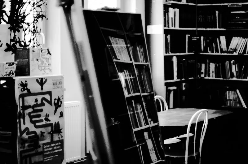 Czarno-białe wnętrze Biblioteki Publicznej w Radwanicach pokazuje regały z książkami, stolik, krzesło i elementy wystawy w eksperymentalnym ujęciu.