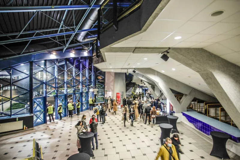 Prostornou halu Vratislavského tréninkového centra Spartan zaplňují účastníci akce, kteří stojí pod ocelovými nosníky u skleněné stěny a tribun.