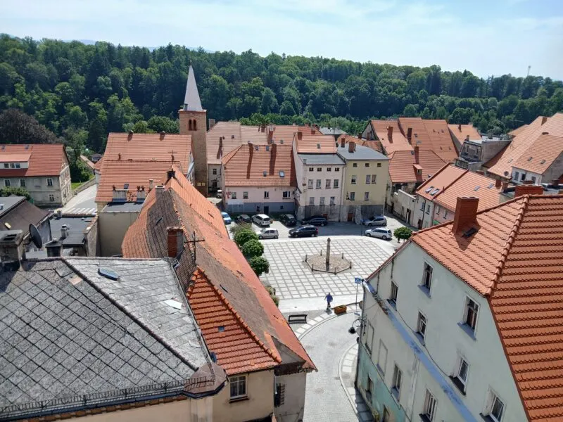 Pohled shora na náměstí s červenými střechami, kostelní věží a zalesněnými kopci v pozadí.