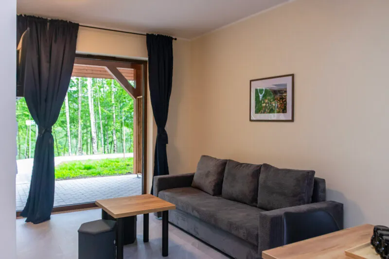 Przestronny apartament z wygodną sofą, stolikiem oraz dużymi drzwiami prowadzącymi na zewnątrz. Duże przeszklenie zapewnia dostęp do naturalnego światła i pozwala cieszyć się widokiem zieleni.