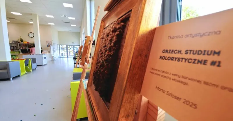 Tkanina artystyczna w brązowej ramie na tle jasnego wnętrza biblioteki, obok opis pracy.