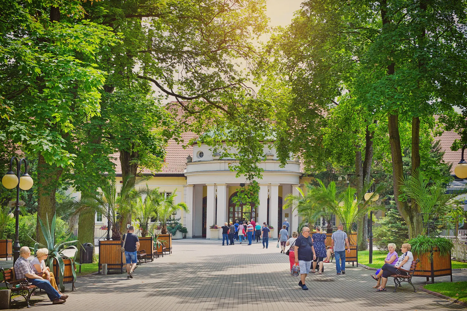 Un ampio viale termale conduce attraverso un parco verde verso un luminoso padiglione, passando accanto a panchine, passanti, alberi secolari e piante ornamentali.