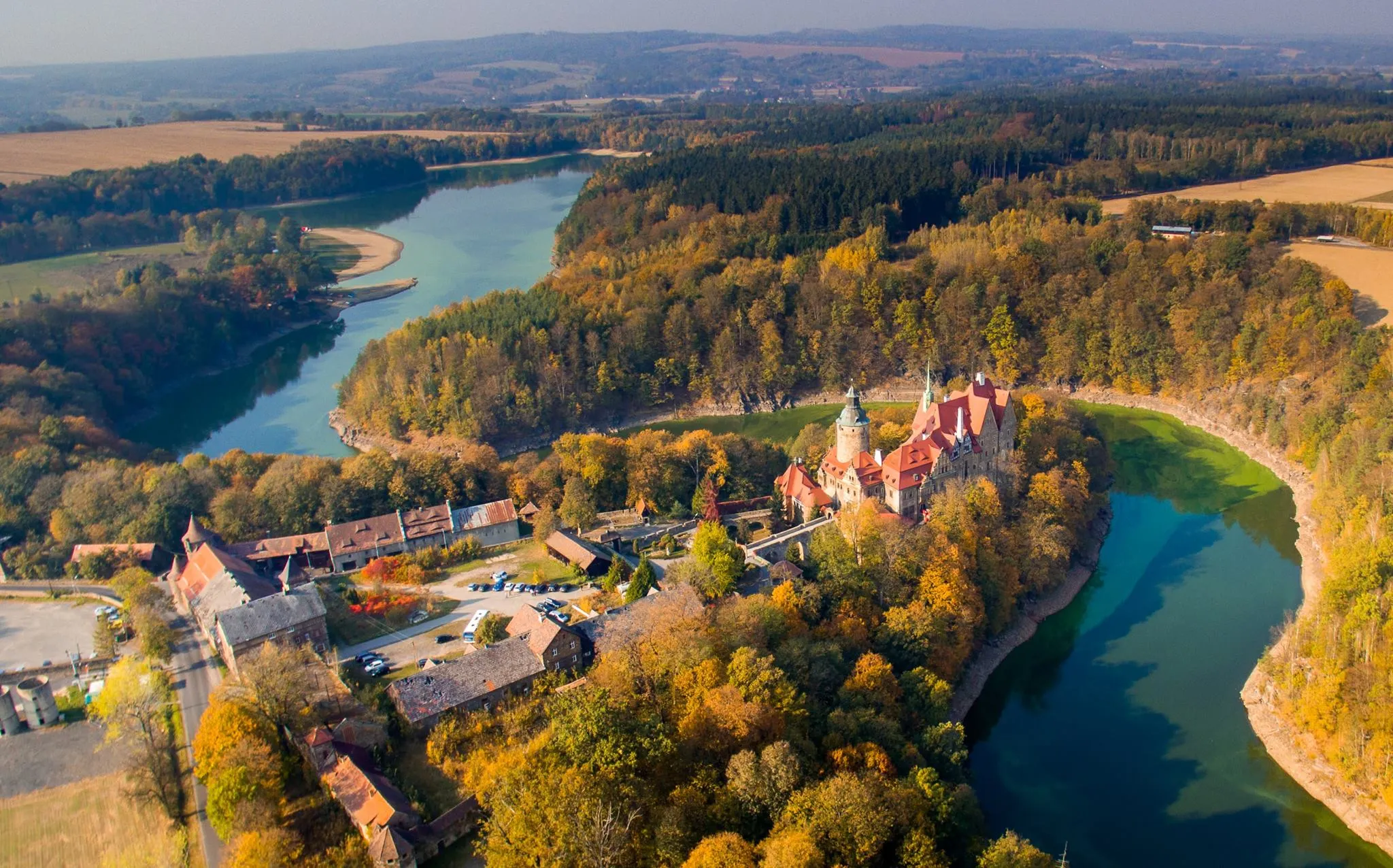 Widok z góry na zamek umiejscowiony nad brzegiem jeziora, które okala las w jesiennych barwach.