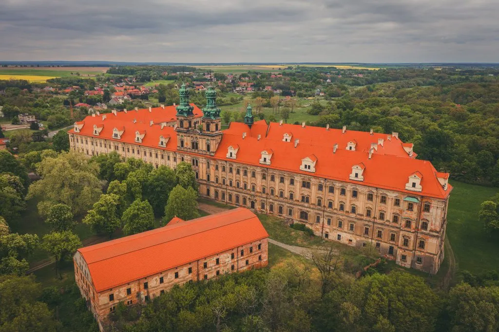 A drone view shows the vast Baroque complex of Lubiąż Abbey with a long facade and red roofs, surrounded by a park, fields, and village buildings.