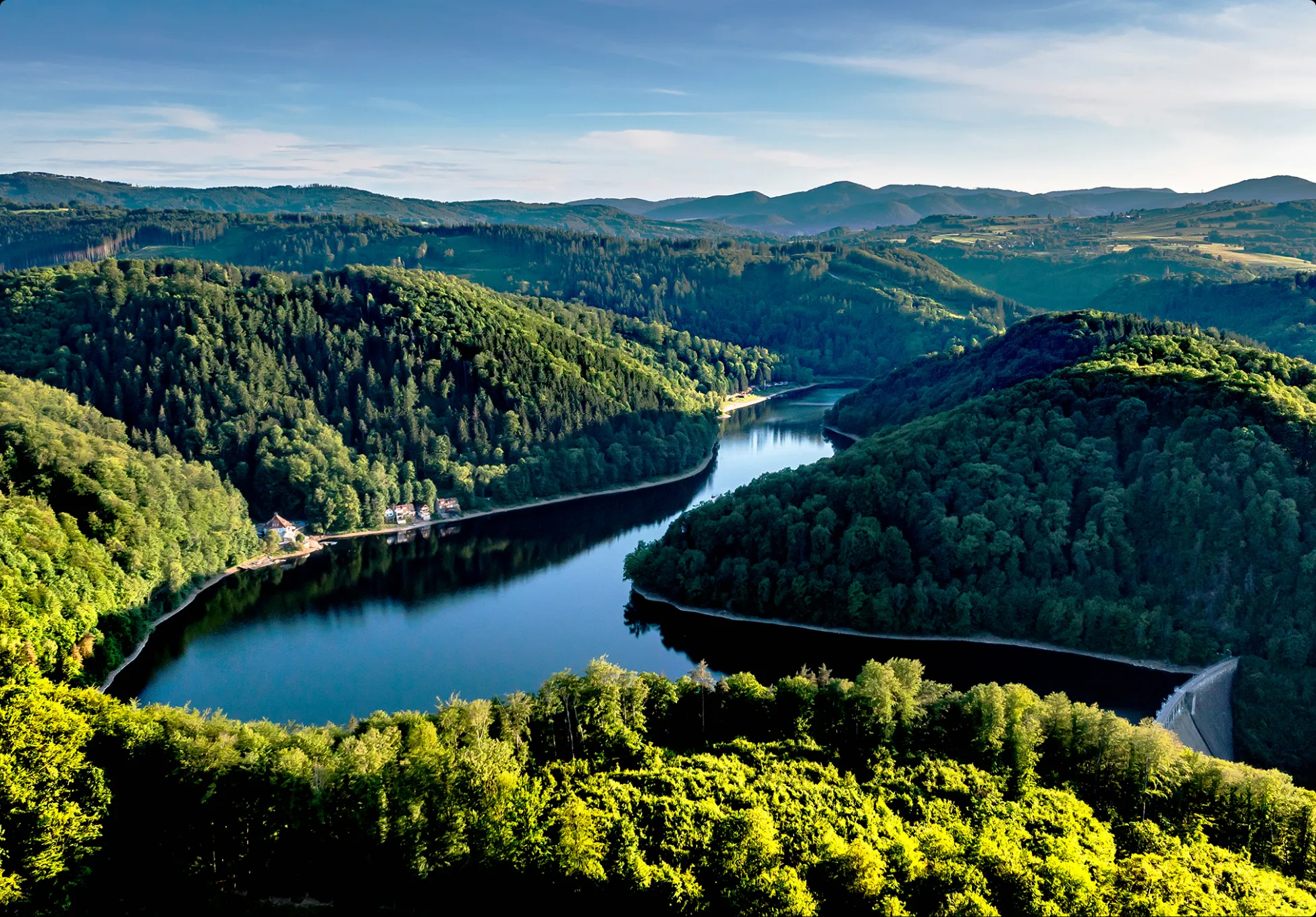 Křivolaká řeka nebo vodní nádrž protíná hustě zalesněné kopce Dolního Slezska a vytváří malebné zákruty na pozadí rozlehlé horské krajiny.