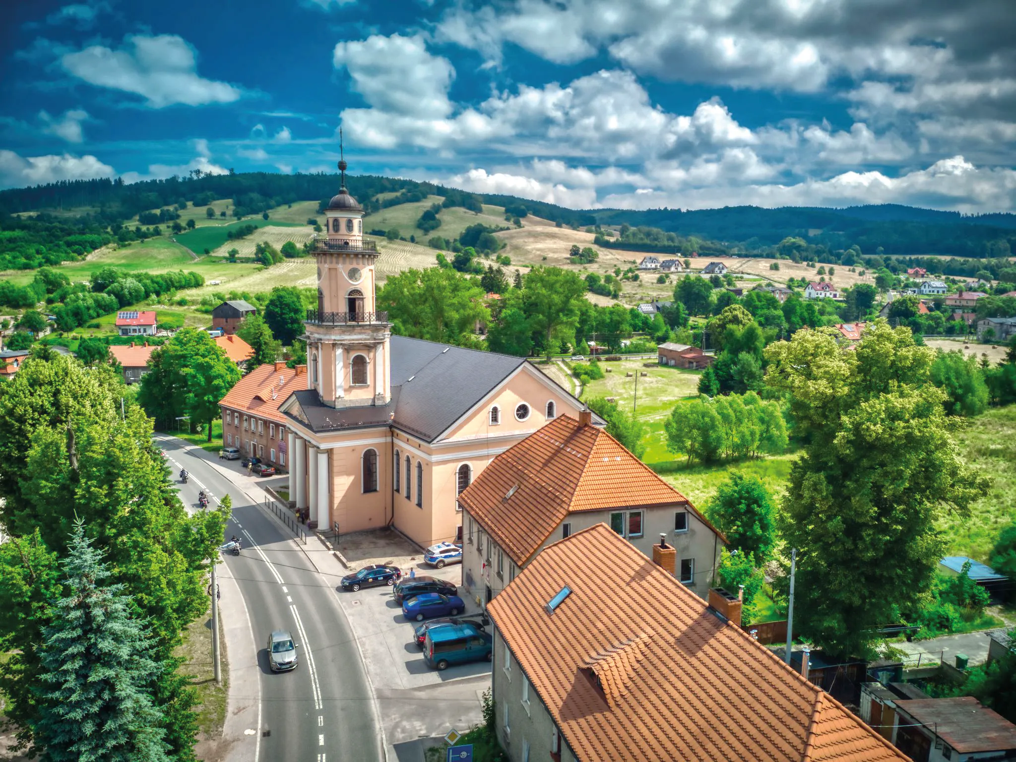 Widok na zabytkowy kościół przy ulicy. Widok na panoramę okolicy Głuszycy Górnej z pofałdowanymi, średniozalesionymi wzniesieniami w tle.