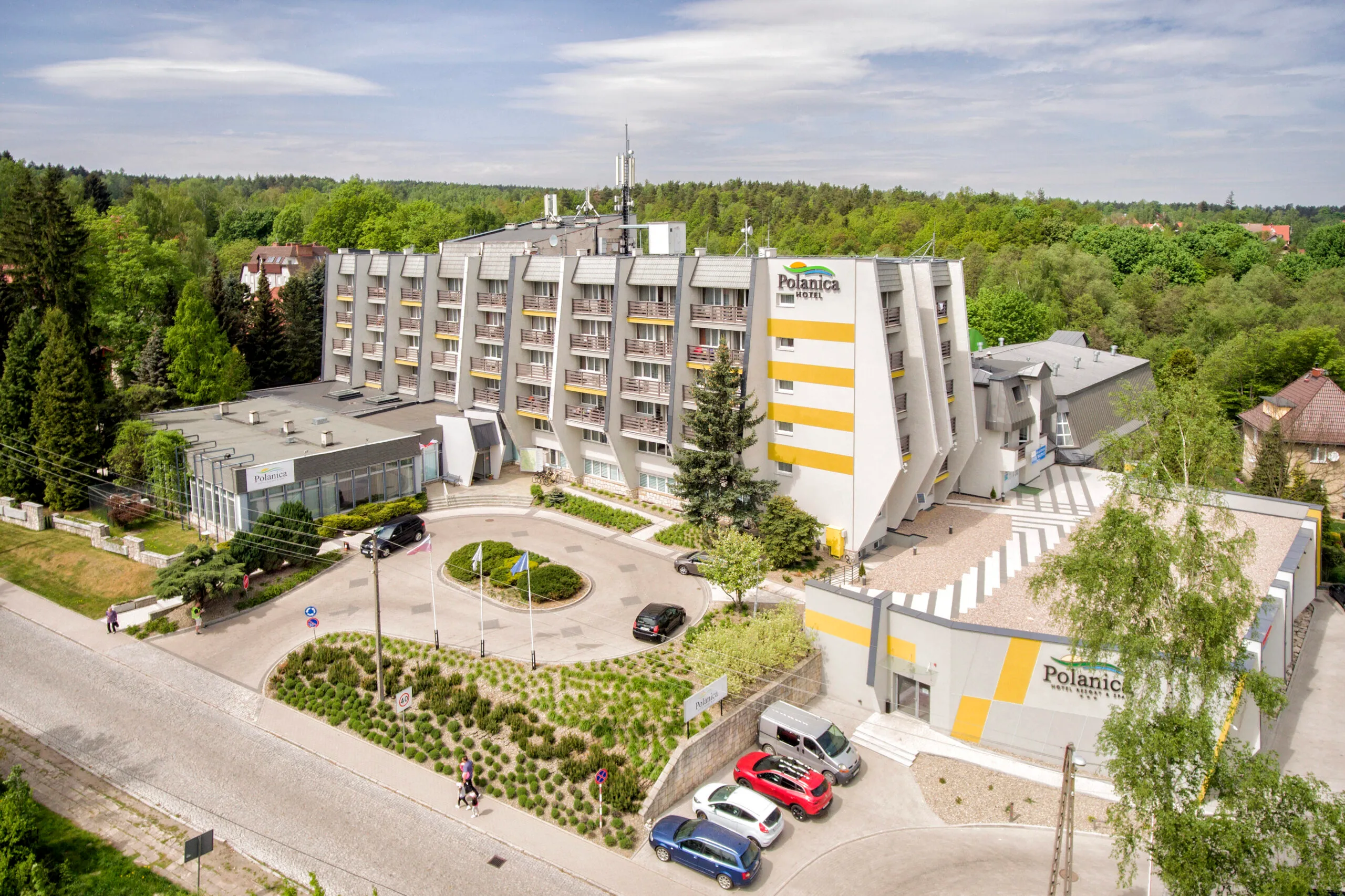 Zdjęcie wykonane z drona, przedstawia masywny, wielopiętrowy budynek hotelowy z podjazdem i parkingiem