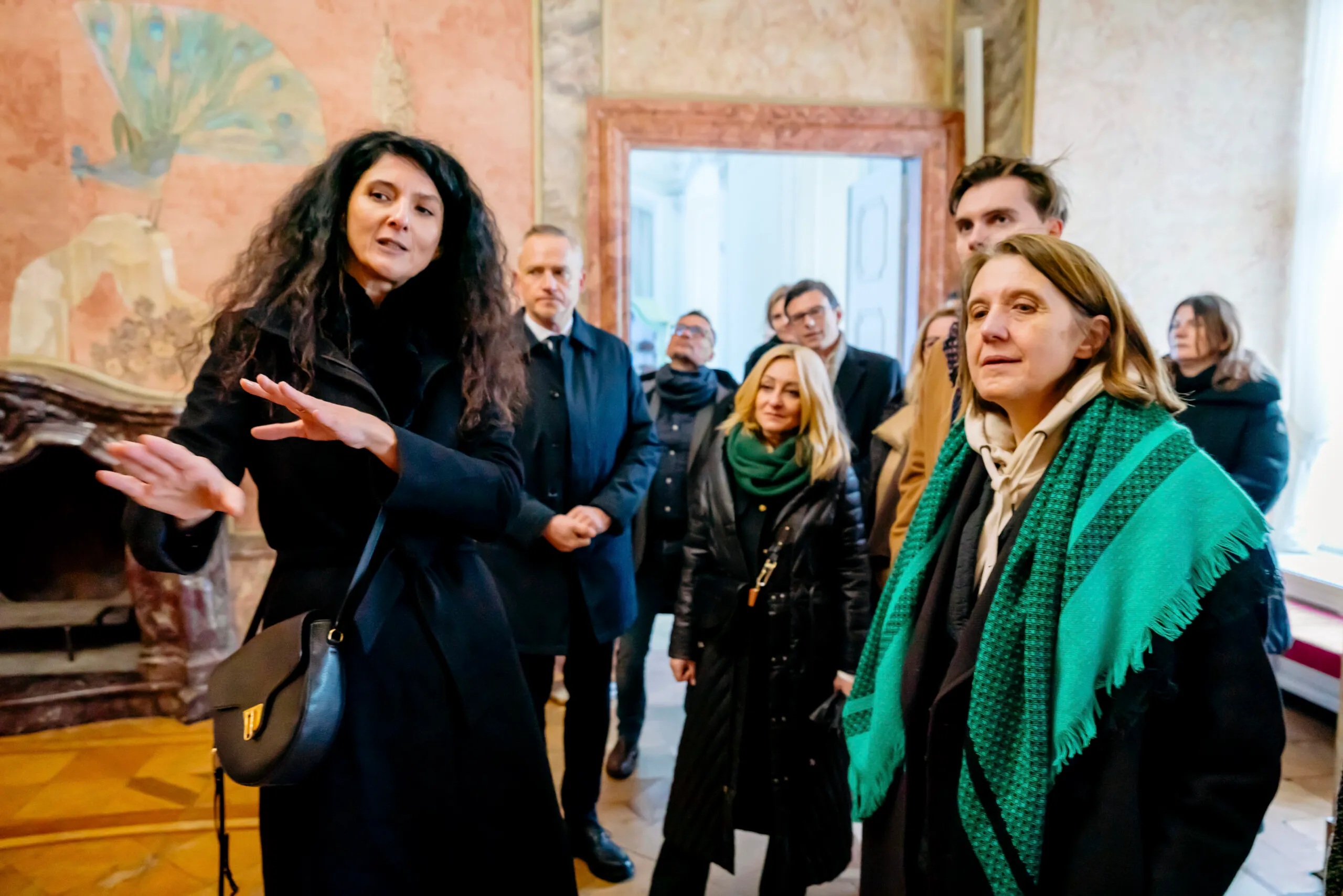 Przewodniczka gestykuluje podczas oprowadzania gości po barokowej sali Zamku Książ, a ministra kultury słucha jej na tle malowideł i sztukaterii.