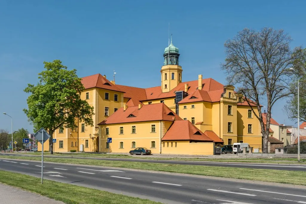 Ceglana, czerwonawo-brązowa budowla z jasnymi detalami wokół okien i bram, wyróżniająca się wieżą w kolorze czerwonej cegły.