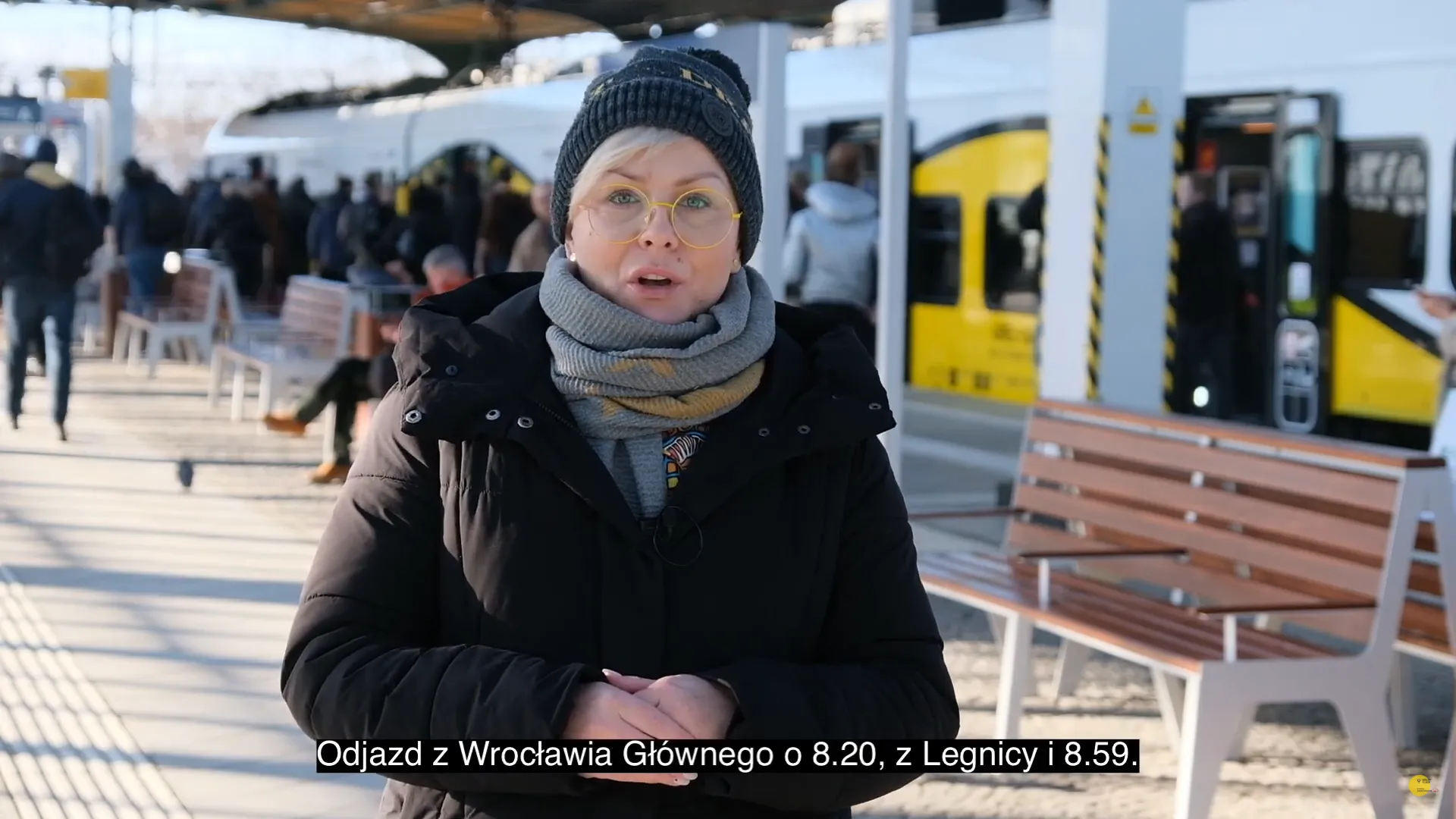 Kobieta w zimowej czapce mówi do kamery na peronie kolejowym, a za nią stoi żółty pociąg i pasażerowie, z podpisem o godzinach odjazdu z Wrocławia i Legnicy.