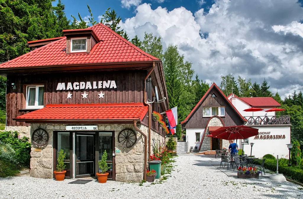 Widok na front budynku dwukondygnacyjnego, obitego drewnem, z kamienną podstawą. Nad wejściem napis Magdalena.