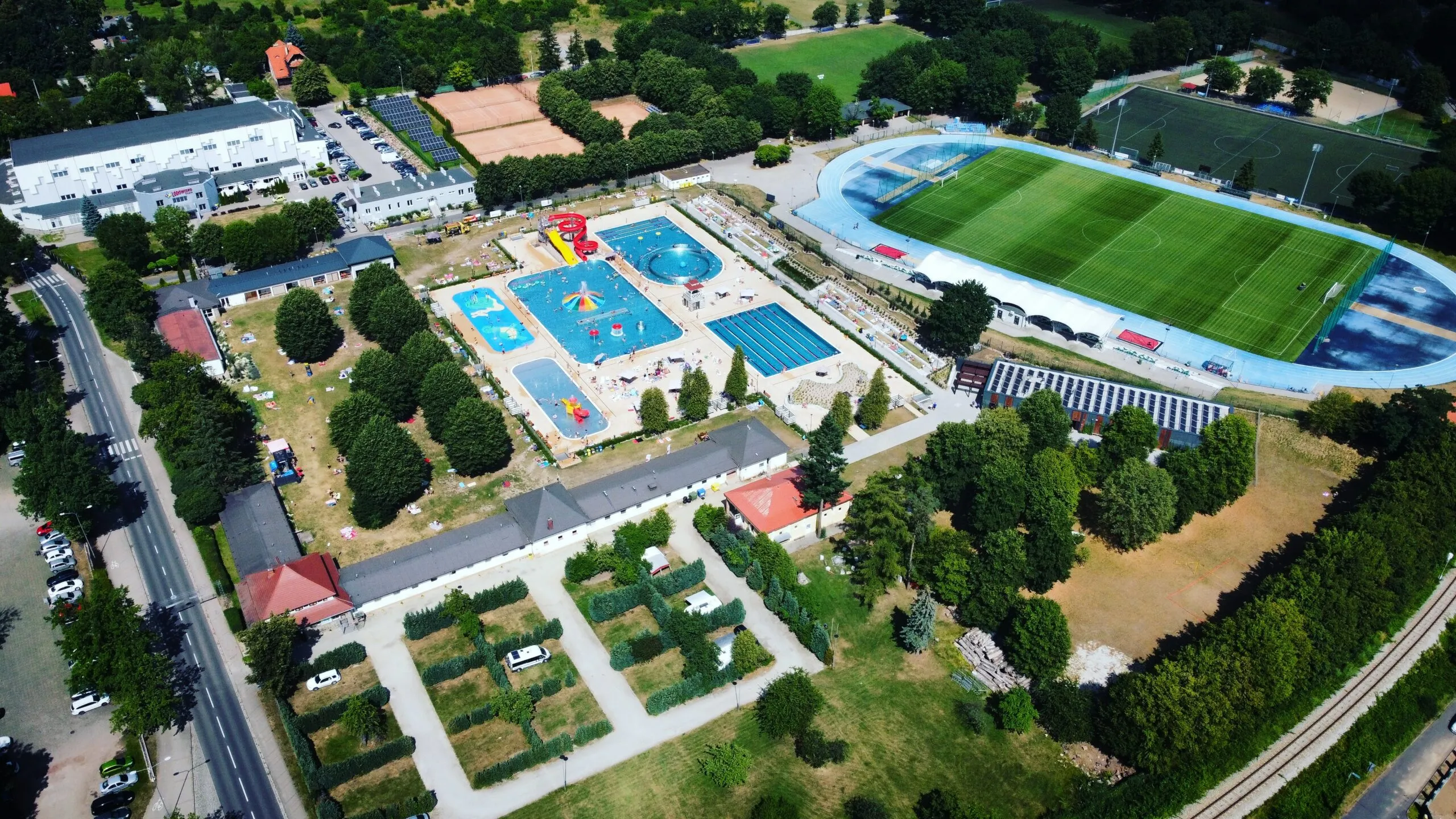 Widok z drona na kompleks sportowo-rekreacyjny przedstawiający między innymi pole campingowe, boisko sportowe oraz baseny zewnętrzne z brodzikiem dla dzieci, podwójną zjeżdżalnią i wirem wodnym. 
