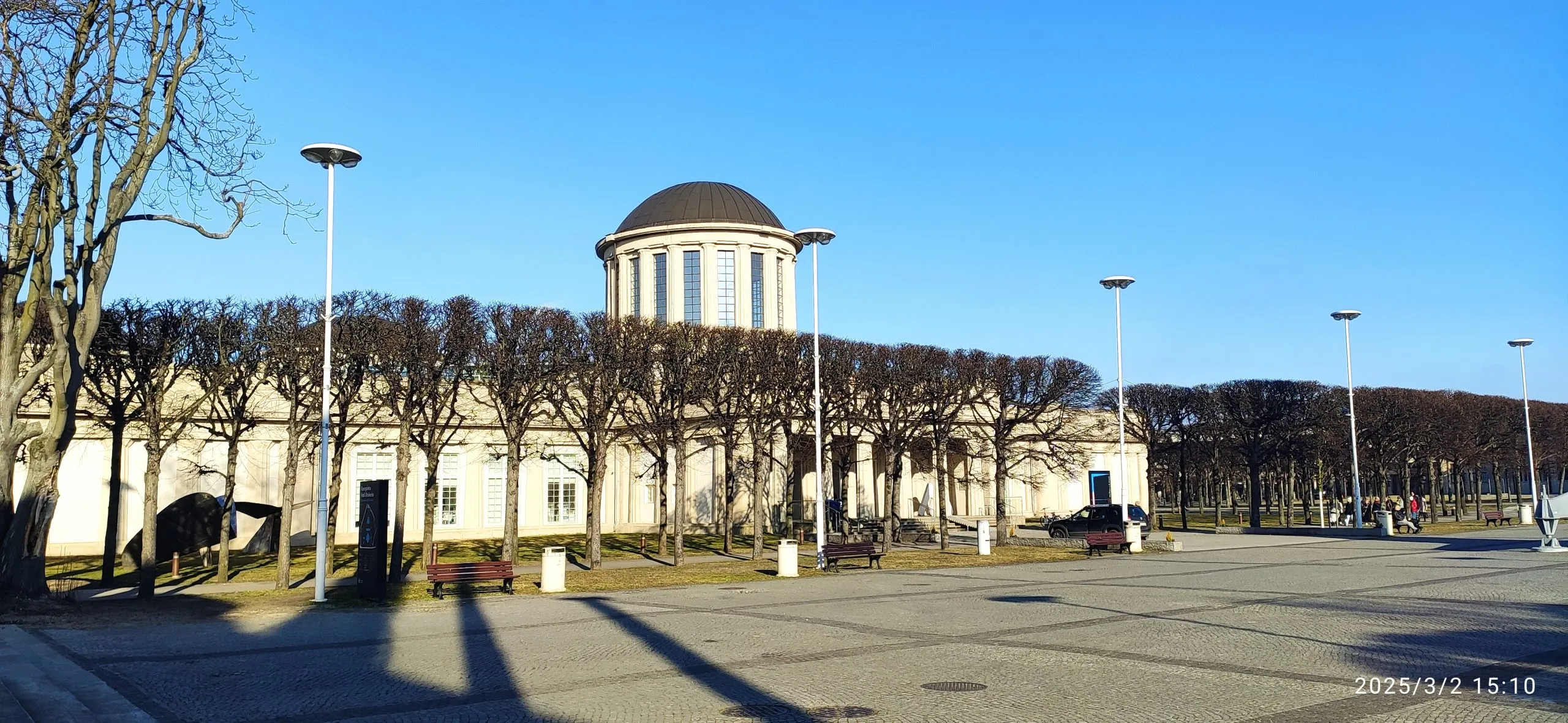 Modernistyczny budynek o prostych, geometrycznych kształtach, wyróżniający się czterema charakterystycznymi kopułami na dachu, otoczony przestronnym terenem.