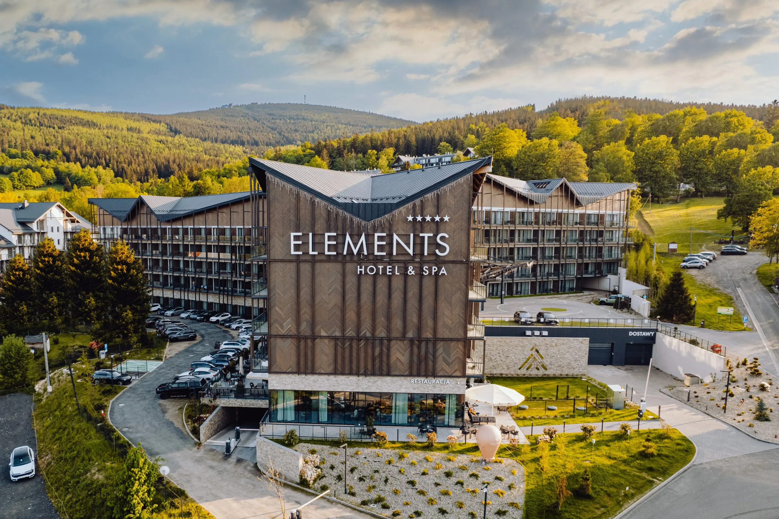 Zdjęcie z drona z dużym budynkiem hotelowym, na szczycie elewacji z elementami drewnianymi logo i nazwa hotelu Elements. Przed budynkiem elementy infrastruktury zielonej, parking. za budynkiem widok na las i góry.
