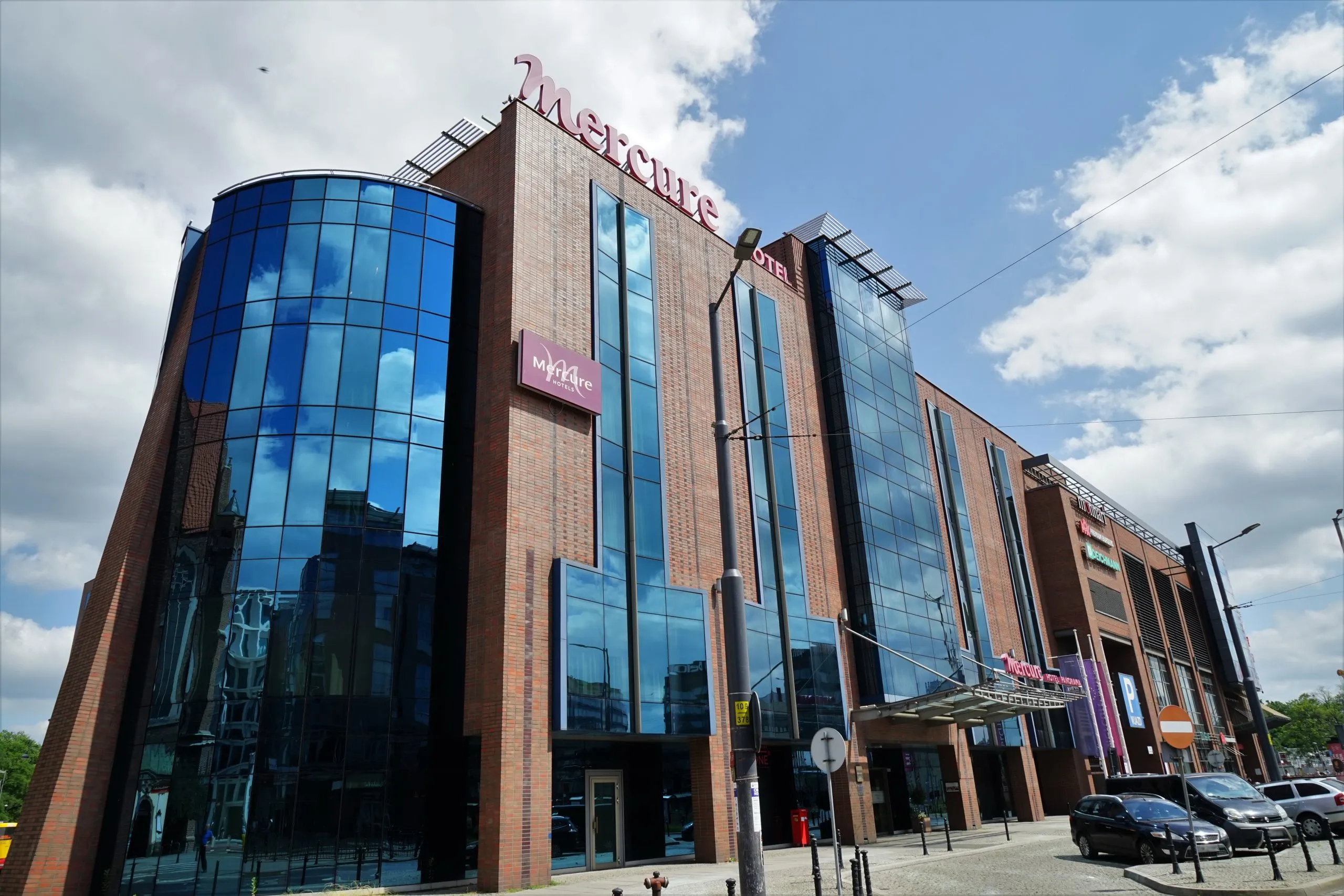 Nowoczesny budynek z fasadą ze szkła oraz materiałów imitujących cegłę. Bryła nieregularna. Na szczycie oraz elewacji logo i nazwa hotelu.