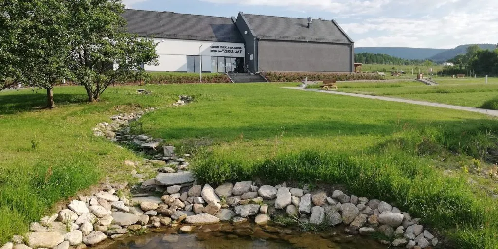 Oczko wodne z kamieniami, za nim zielona łąka oraz nowoczesny parterowy budynek centrum edukacyjnego