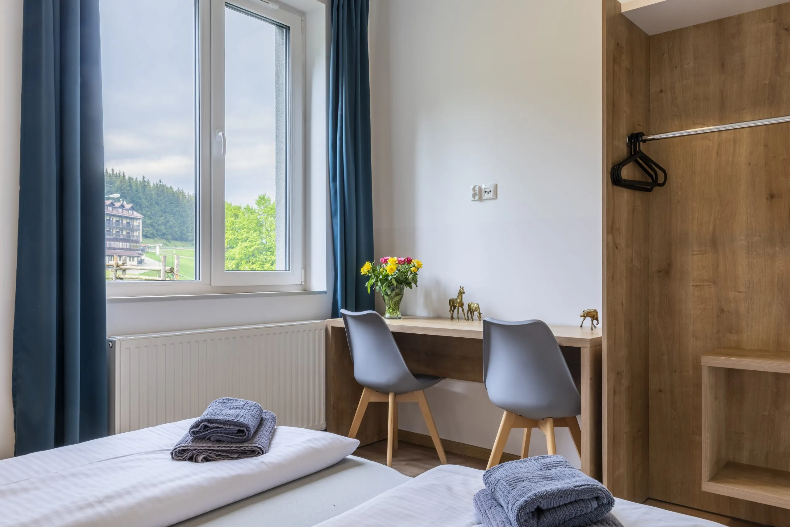Nowoczesny pokój hotelowy z dwoma łóżkami, biurkiem, dużą szafą oraz dużym oknem, przez które rozpościera się widok na górski krajobraz. Wnętrze jest utrzymane w naturalnych, ciepłych barwach, z drewnianymi elementami i minimalistycznymi dekoracjami, takimi jak złote figurki zwierząt na biurku. Na łóżkach złożone ręczniki.