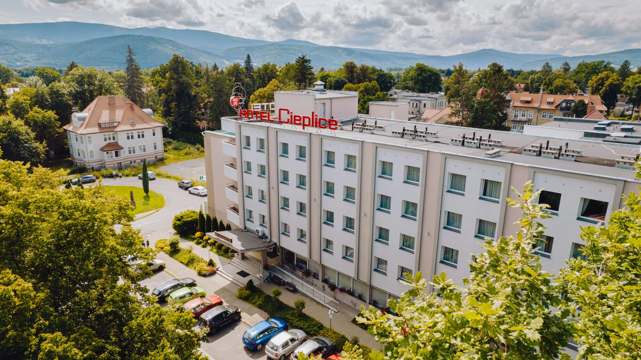 Duży prostokątny budynek z napisem "Hotel Cieplice" na szczycie fasady, na tle zieleni oraz gór.