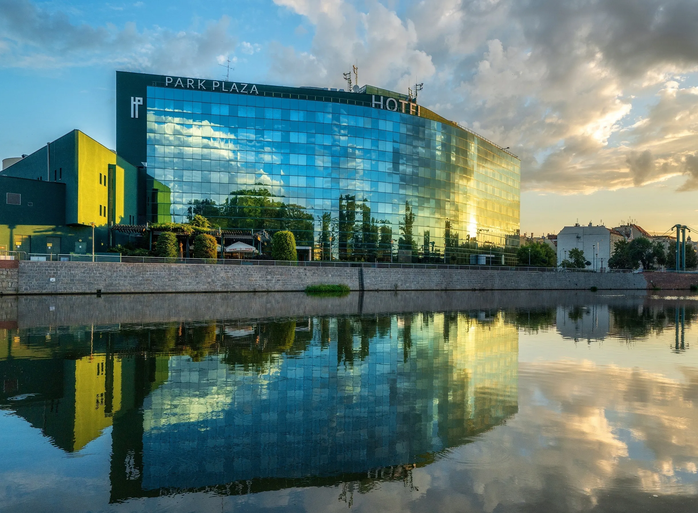 Duży szklany prostokątny budynek, zlokalizowany nad rzeką, Na szczycie nazwa oraz logo hotelu: "HP Hotel Plaza".