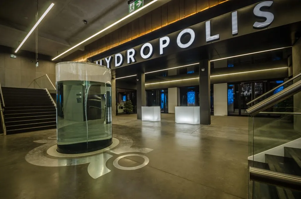 Interiér moderní budovy – v popředí průhledný skleněný válec s vířící vodou, v pozadí nápis „Hydropolis“