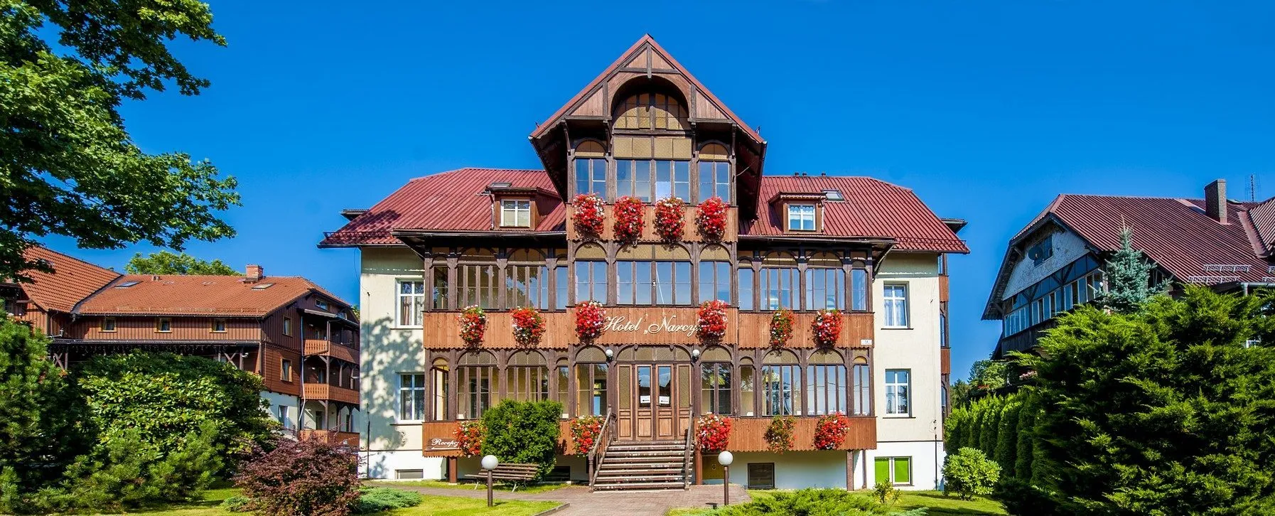 Hotel od frontu, z dobrze widocznym wejściem i otaczającym go terenem. Jasna kolorystyka budynku i duże okna podkreślają jego elegancki, uzdrowiskowy styl.