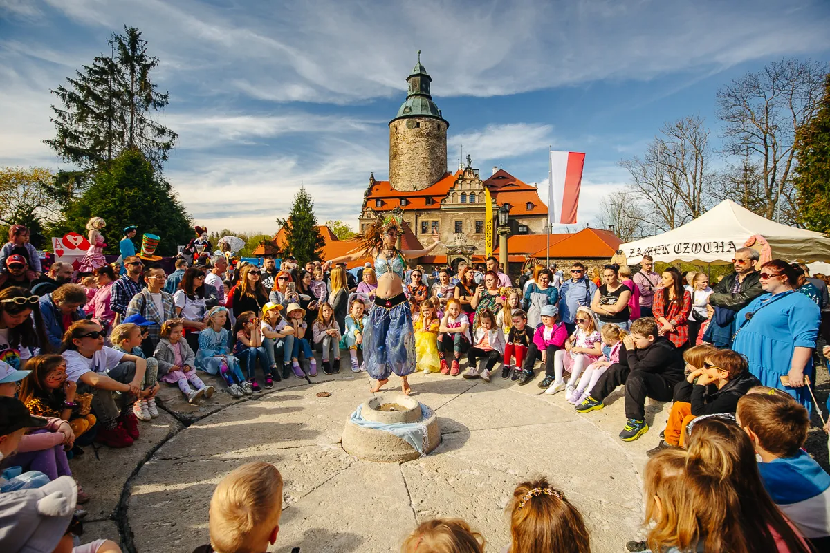 Duża grupa uczestników pozuje wokół rzeźby na placu przed zamkiem z wysoką wieżą, podczas plenerowego spotkania w słoneczny dzień.