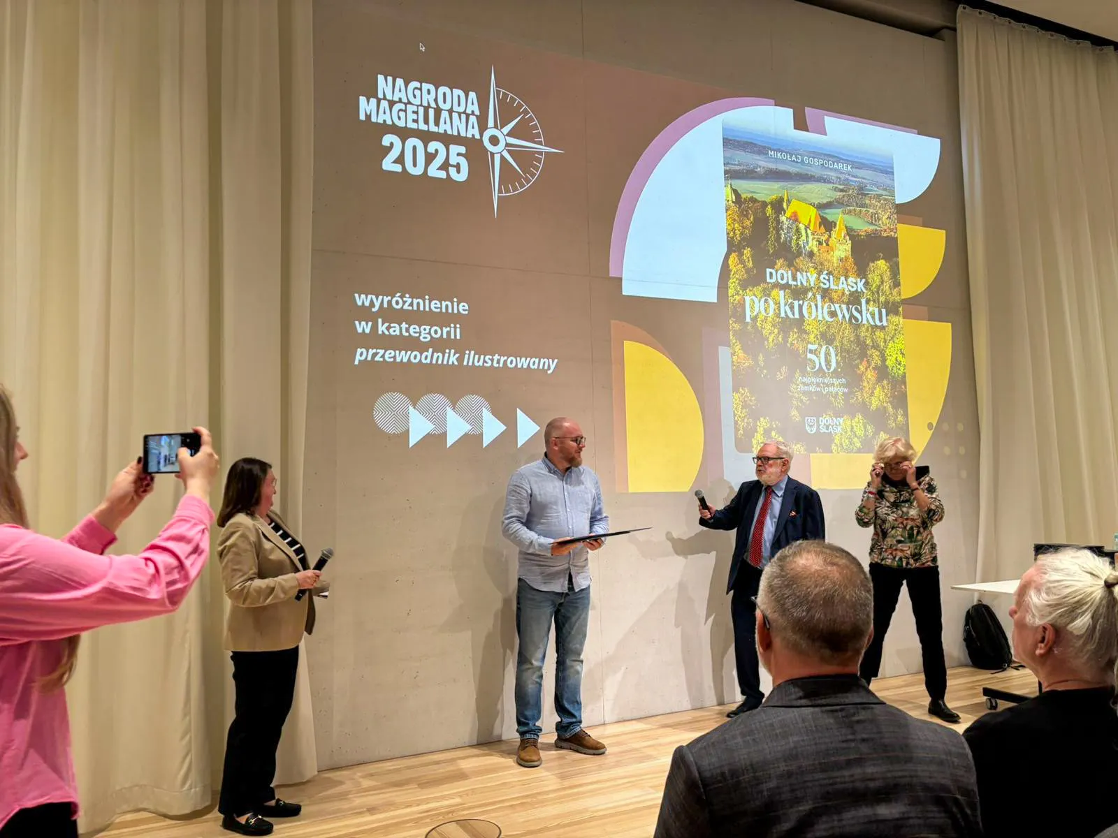 Mężczyzna prezentuje nagrodzoną publikację „Dolny Śląsk po królewsku” podczas uroczystości, stojąc na scenie przed publicznością i ekranem z wyróżnieniem.