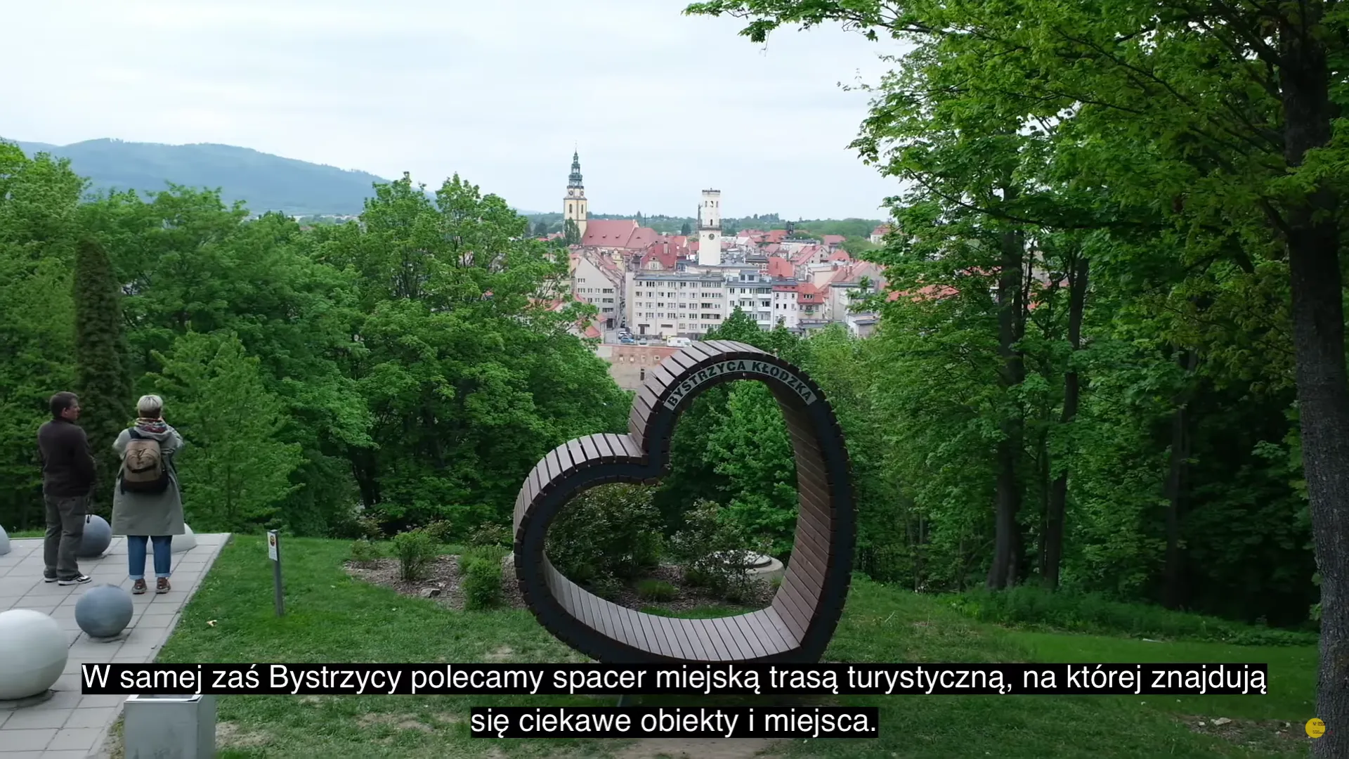 Kadr z filmu o Bystrzycy Kłodzkiej pokazuje parkową instalację z dużymi metalowymi okręgami oraz panoramę miasta widoczną między zielonymi drzewami.