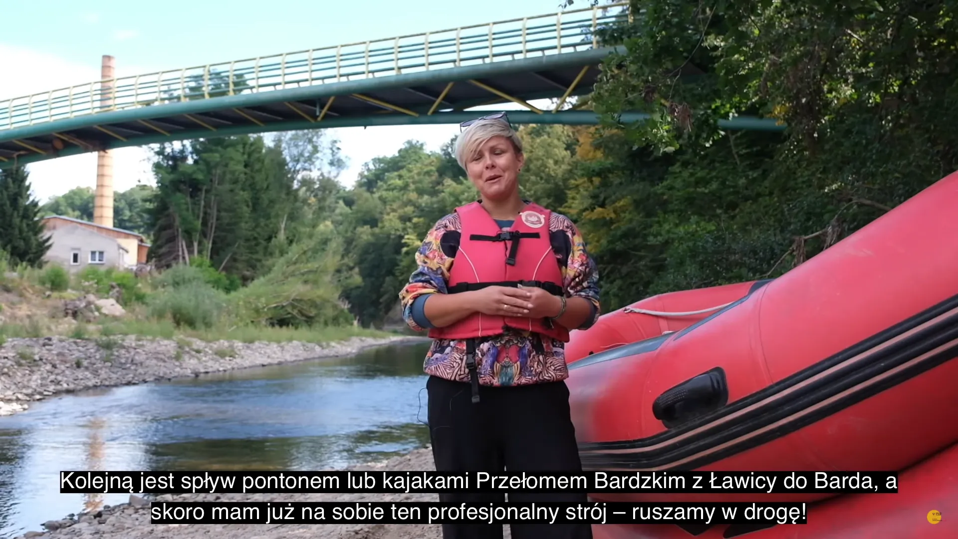 Kobieta w kamizelce asekuracyjnej opowiada do kamery przed czerwonym pontonem nad Przełomem Bardzkim, zapowiadając spływ do Barda.