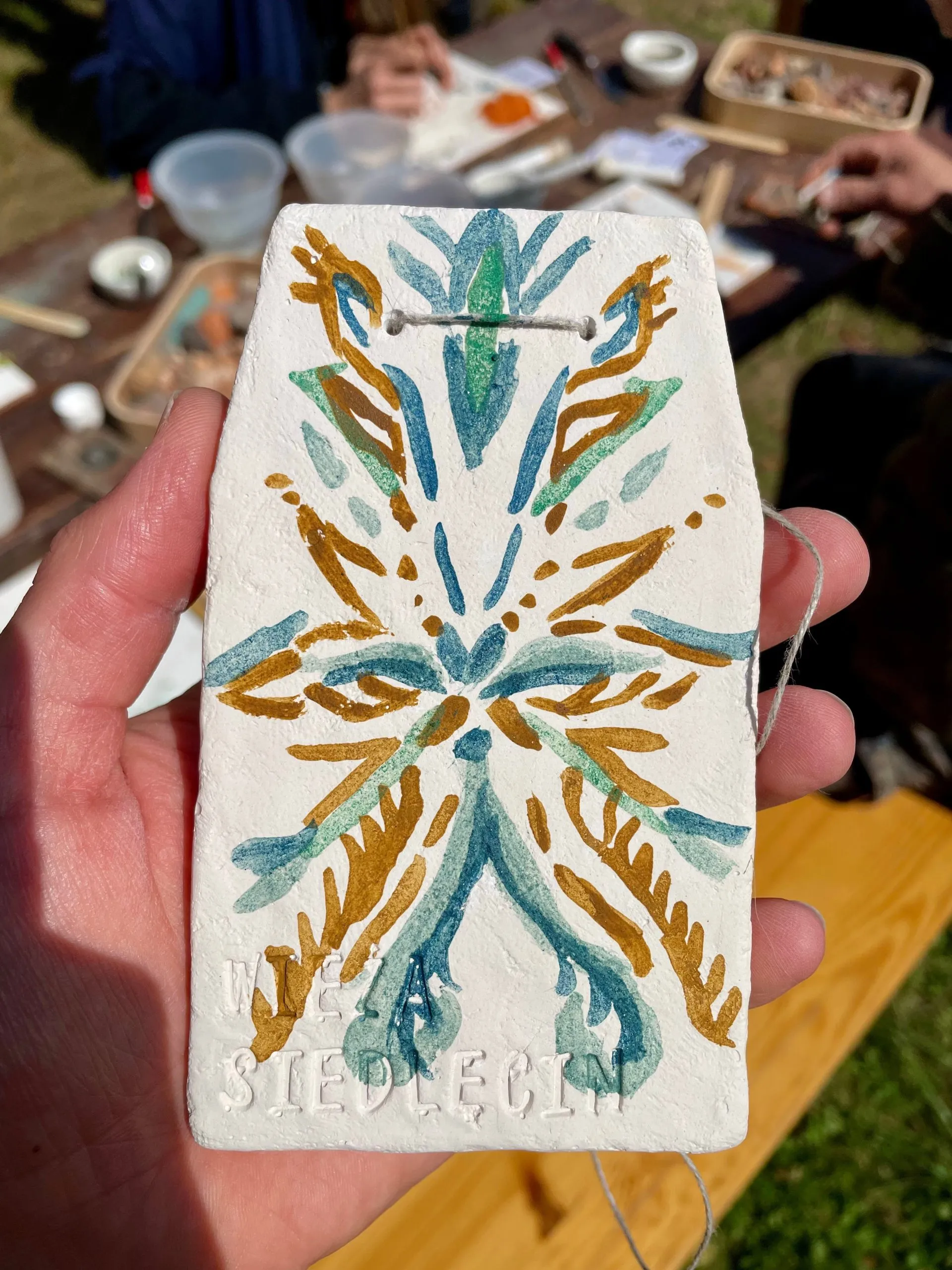 Dłoń trzyma ceramiczną płytkę ozdobioną podczas warsztatów pigmentów przy Wieży Siedlęcin, z turkusowym i ochrowym wzorem przypominającym liście.